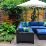 A beautiful backyard containing green plants, bright flowers, a blue couch, a fence, and a tall umbrella.