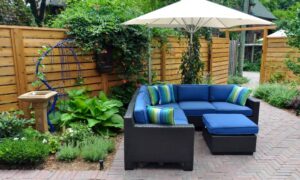 A beautiful backyard containing green plants, bright flowers, a blue couch, a fence, and a tall umbrella.