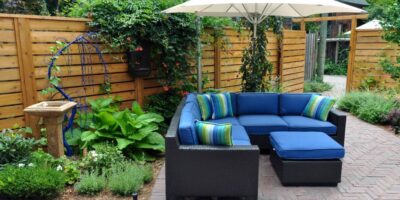 A beautiful backyard containing green plants, bright flowers, a blue couch, a fence, and a tall umbrella.