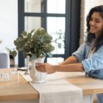 A smiling young woman setting a vase containing a broad-leafed plant on top of a wooden dining table.