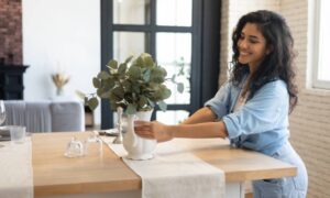 A smiling young woman setting a vase containing a broad-leafed plant on top of a wooden dining table.