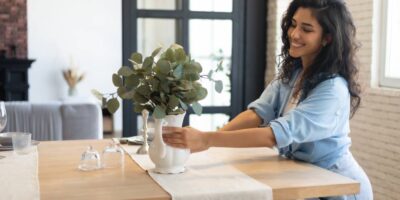 A smiling young woman setting a vase containing a broad-leafed plant on top of a wooden dining table.