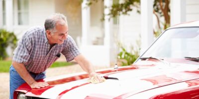 An older man is washing his vintage red luxury sports car with white stripes. He's applying wax to the hood.