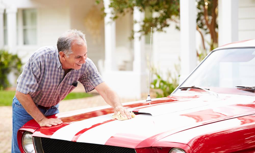 An older man is washing his vintage red luxury sports car with white stripes. He's applying wax to the hood.