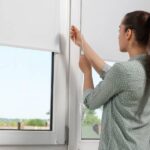 A woman with a blouse and ponytail adjusts her roller blinds on her home windows overlooking her neighborhood.