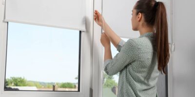 A woman with a blouse and ponytail adjusts her roller blinds on her home windows overlooking her neighborhood.