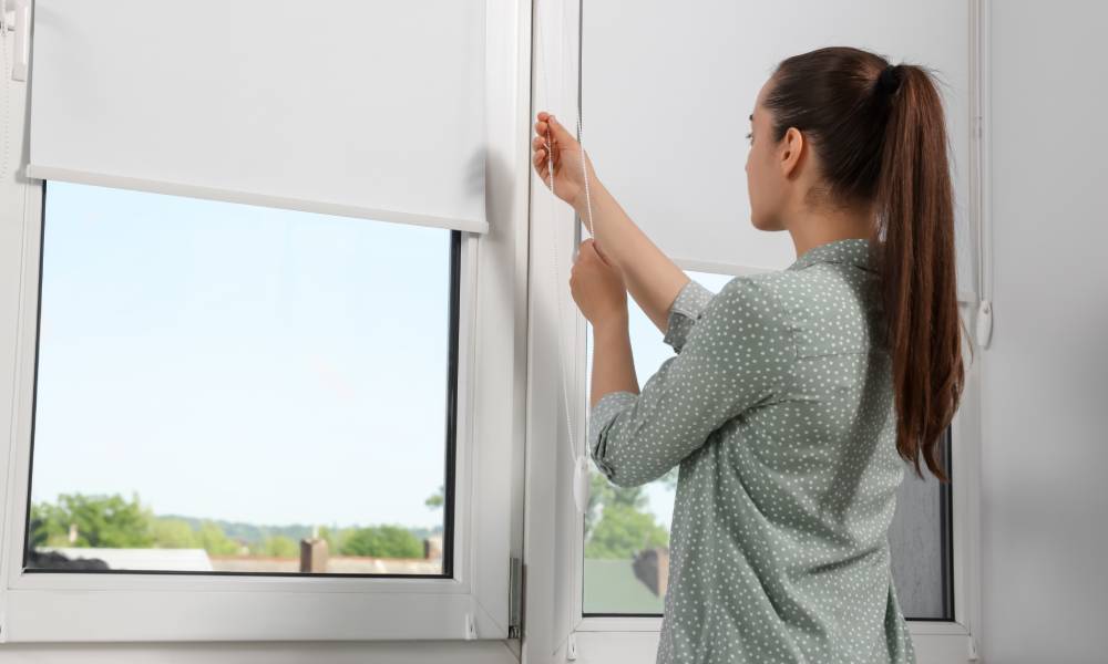 A woman with a blouse and ponytail adjusts her roller blinds on her home windows overlooking her neighborhood.