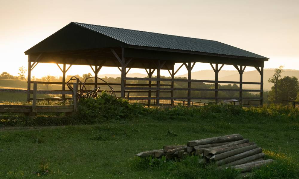 An old pole barn is in front of a fence and a pile of logs with the sun and mountains in the background.