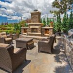 An outdoor living space lies before a natural background. The center has a seating area with a grill and fireplace to the side.