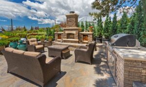 An outdoor living space lies before a natural background. The center has a seating area with a grill and fireplace to the side.