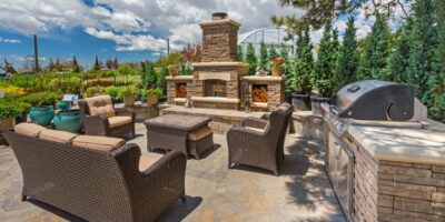 An outdoor living space lies before a natural background. The center has a seating area with a grill and fireplace to the side.