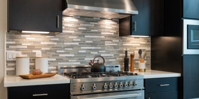 A modern kitchen with a stainless steel gas range and hood. The countertops are white and the cabinets are black.