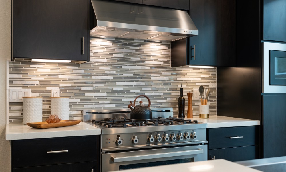 A modern kitchen with a stainless steel gas range and hood. The countertops are white and the cabinets are black.