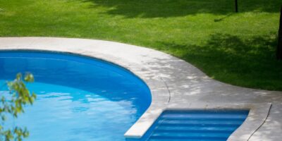 A concrete pool deck surrounds a beautiful, idyllic inground pool. Bright, green grass surrounds the pool deck.