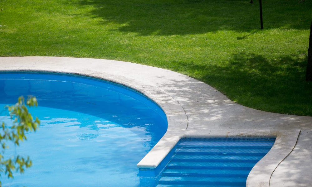 A concrete pool deck surrounds a beautiful, idyllic inground pool. Bright, green grass surrounds the pool deck.