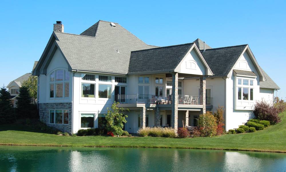 A beautiful luxury home sits on the edge of a lakefront property with gorgeous landscaping and blue skies in the background.