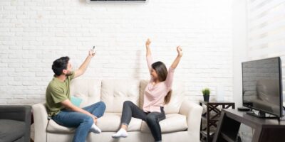 A man and woman sit on a couch. The man uses a remote to control a mini split above them while the woman cheers.