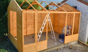 An older man wearing a jacket and jeans kneeling down with a power tool while working on a wooden building.