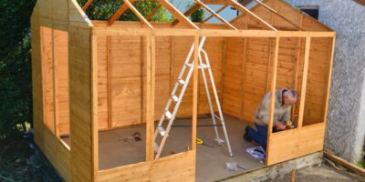 An older man wearing a jacket and jeans kneeling down with a power tool while working on a wooden building.