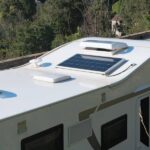 An overhead view of an RV parked on a roadway in full sun. The RV's roof has a solar panel and the RV roof vents are open.