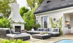 A luxury backyard patio with matching grey wicker furniture and a white stone fireplace leading to an indoor kitchen.