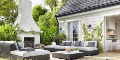 A luxury backyard patio with matching grey wicker furniture and a white stone fireplace leading to an indoor kitchen.