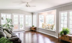 A sunroom with gray, paneled walls, a brown leather couch, and a window seat with pillows sitting on it.