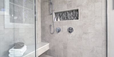 A gray-tiled walk-in shower contains a bench with a white towel and a gray washcloth. There's a window in the shower.