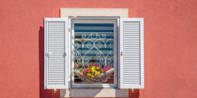 Close-up of a window on a coral-colored building with white open shutters and a flower basket with yellow and pink daisies.