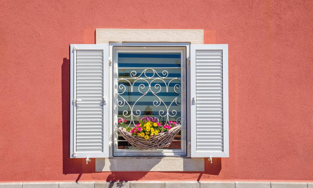 Close-up of a window on a coral-colored building with white open shutters and a flower basket with yellow and pink daisies.