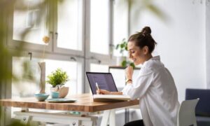 A woman sits at a wooden desk in front of large, picturesque windows. A laptop is open, and she's writing in a notebook.