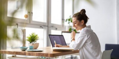 A woman sits at a wooden desk in front of large, picturesque windows. A laptop is open, and she's writing in a notebook.