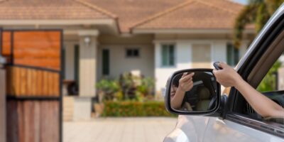 A person driving a silver car pushes the button on a remote that opens an automatic gate for their home's driveway.