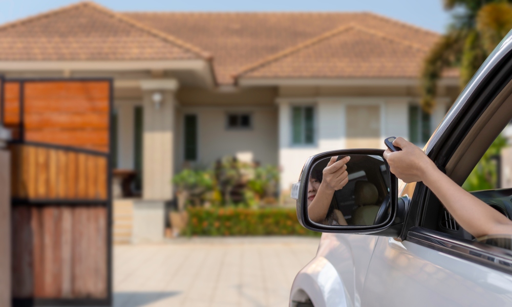 A person driving a silver car pushes the button on a remote that opens an automatic gate for their home's driveway.