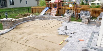 A patio area of a fenced-in yard remains partially completed as sand and stone bricks cover portions of the area.