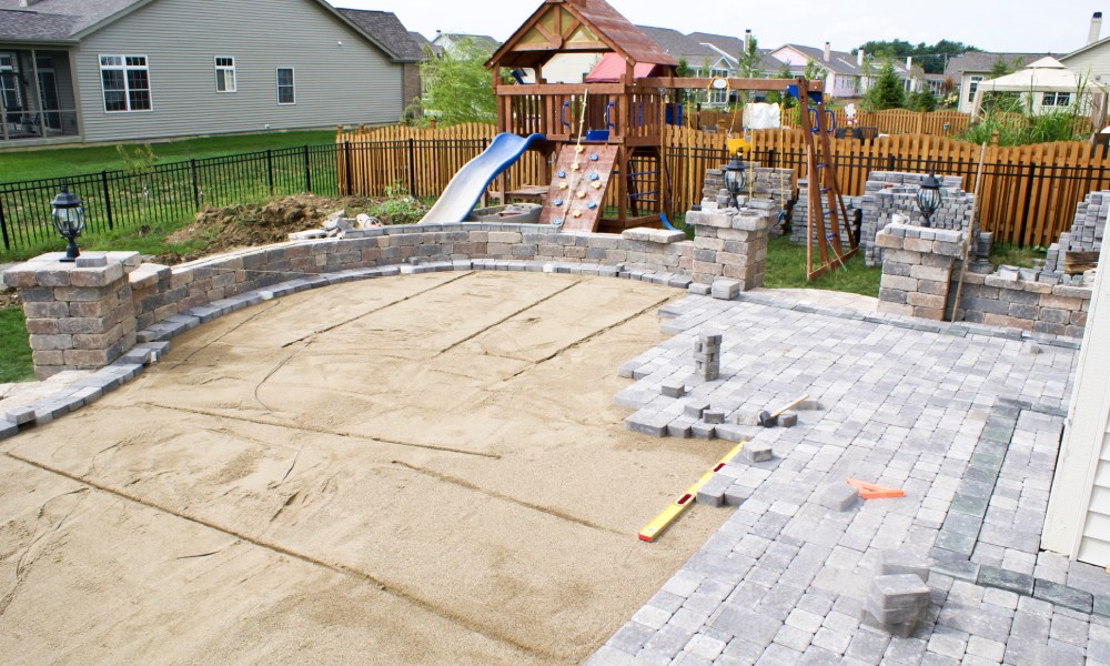 A patio area of a fenced-in yard remains partially completed as sand and stone bricks cover portions of the area.