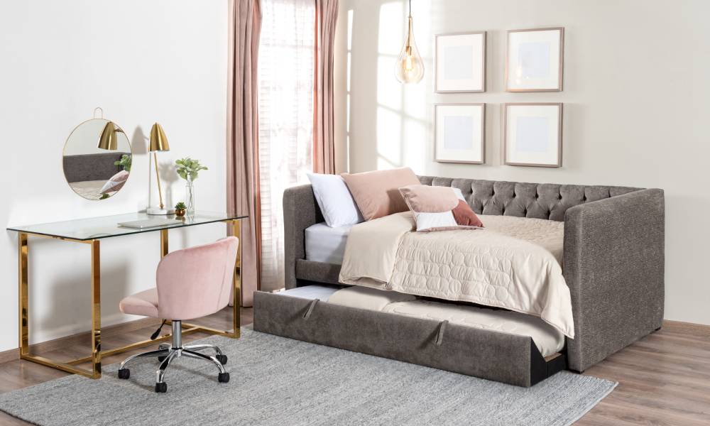 A room with an office desk and a day bed near the window. The furniture has a soft tone color of pink, white, tan, and gray.