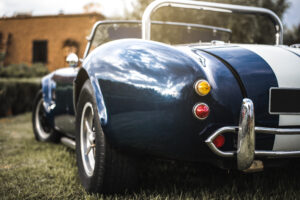 A rear view of a blue vintage convertible with white racing stripes, chrome accents, and round taillights parked on grass.