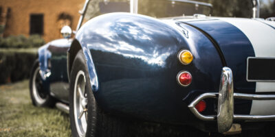 A rear view of a blue vintage convertible with white racing stripes, chrome accents, and round taillights parked on grass.