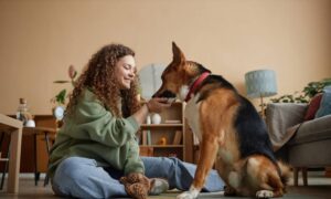 A smiling young woman sits on the living room floor feeding her dog a treat. A mirror and a bookshelf are in the background.