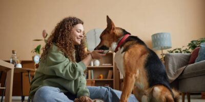 A smiling young woman sits on the living room floor feeding her dog a treat. A mirror and a bookshelf are in the background.