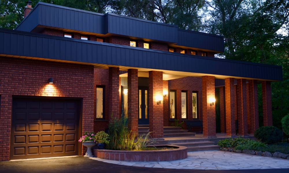 Front of a red brick home at twilight with a pond and stone porch. Lamps illuminate the flat roof house with trees in the background.