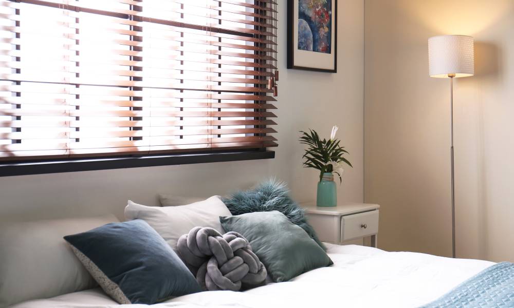 A bedroom with a large window, a made bed with teal bedding, a side table with flowers, artwork, and a floor lamp.