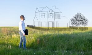 A man is standing in the middle of a field, imagining a newly built, two-story home with a tree planted next to it.