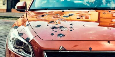 The hood of a red car covered in bird droppings. There are also some bird droppings on the windshield of the car.