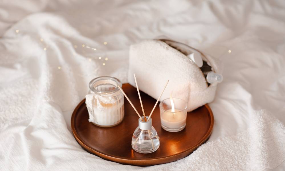 Several scented objects, including two candles and a reed diffuser, on a metallic plate. The plate is on a white sheet.