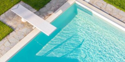 An aerial view of a rectangular swimming pool surrounded by a green lawn. A white diving board extends over the blue water.