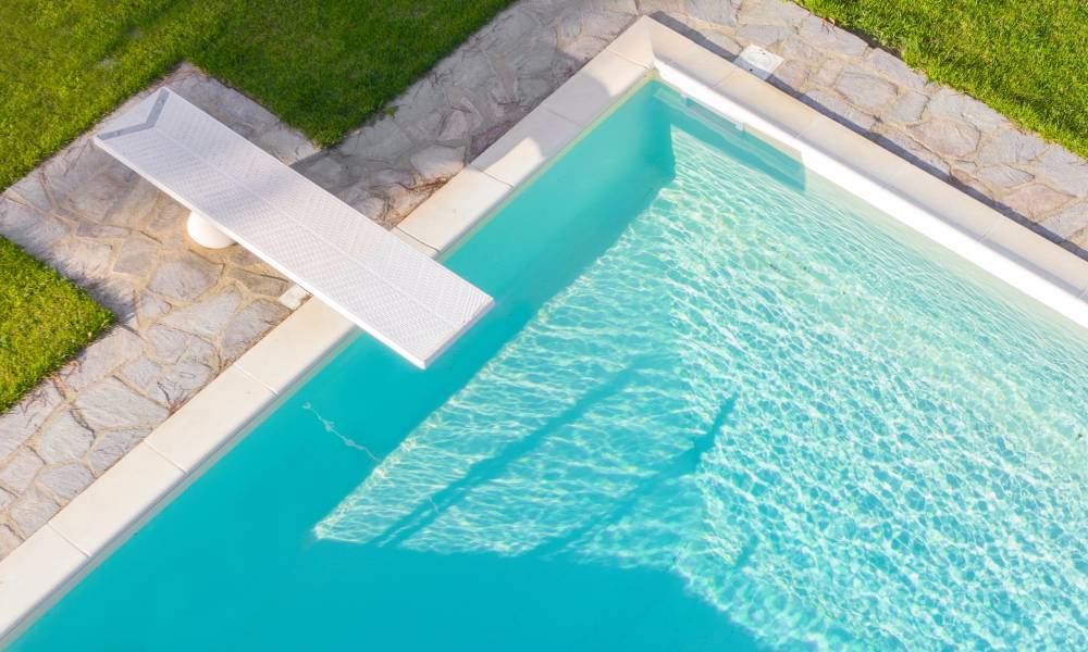 An aerial view of a rectangular swimming pool surrounded by a green lawn. A white diving board extends over the blue water.