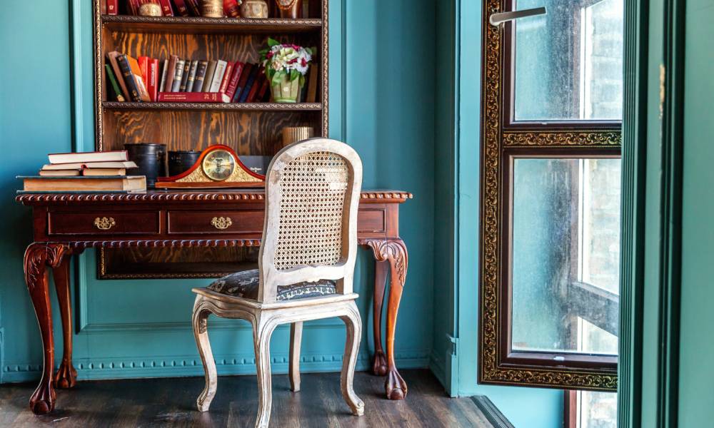 The corner of a room near an open window with an old wooden desk, chair, and bookshelf built into the green wall.