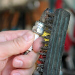 A person using a metal brush to clean a dirty spark plug. The background is out of focus but appears to be a garage.